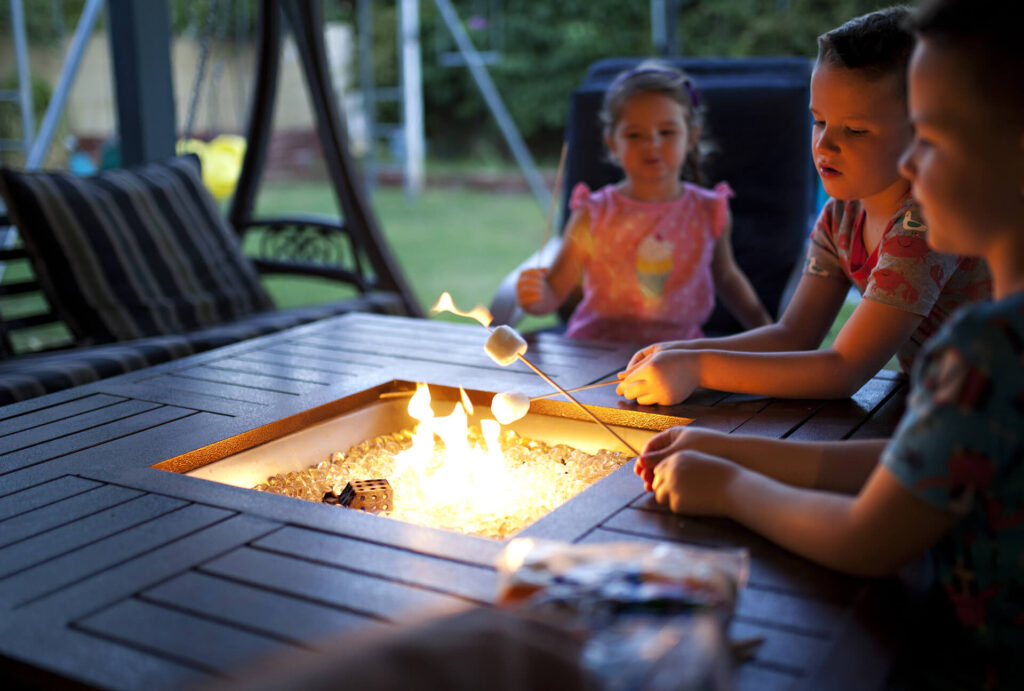 A close-up image of a square shaped black metal patio fire pit with decorative sides, filled with burning logs and orange-yellow flames. Ideal for outdoor gatherings and creating a warm atmosphere. Masonry professionals and turf installers.